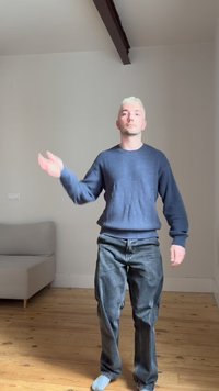 Mladý muž s krátkymi blond vlasmi v modrom svetri a džínsoch, stojaci bosý na drevenej podlahe, gestikulujúci ľavou rukou v minimalistickej miestnosti.