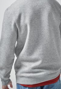 Grauer Pullover aus weichem Stoff, mit Rundhalsausschnitt und gerippten Bündchen. Wird über einem roten Hemd getragen, mit lässiger Passform. Rückansicht sichtbar.