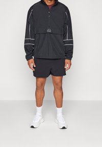 Mężczyzna ubrany w czarną wiatrówkę z białymi lamówkami, czarne krótkie spodenki, białe skarpety i białe trampki, stoi na jednolitym jasnym tle.