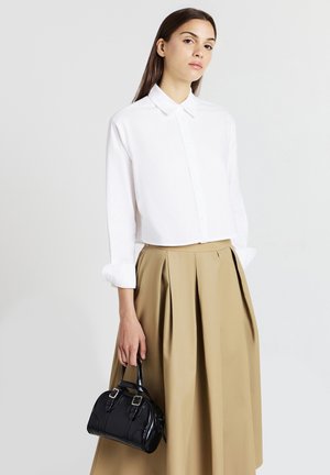 Donna che indossa una camicia bianca con bottoni e una gonna plissettata beige, mentre tiene una piccola borsa nera su uno sfondo semplice.