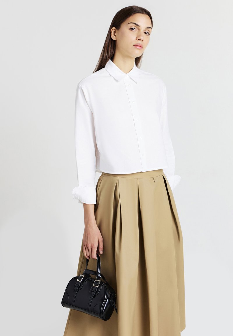 Donna che indossa una camicia bianca con bottoni e una gonna plissettata beige, mentre tiene una piccola borsa nera su uno sfondo semplice.