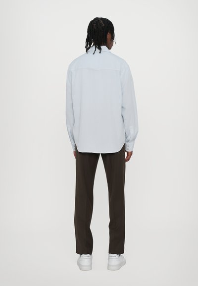 Svetlo modrá košeľa s golierom, dlhými rukávmi a gombíkmi na manžetách, v kombinácii s hnedými nohavicami a bielymi teniskami.