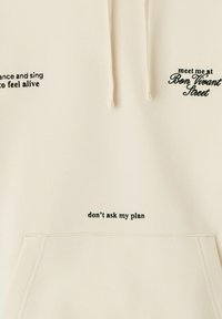 Sweat-shirt couleur crème avec du texte noir brodé. Il est doté d'une capuche avec cordons et d'une poche frontale. Le texte comprend des phrases et des détails de localisation.