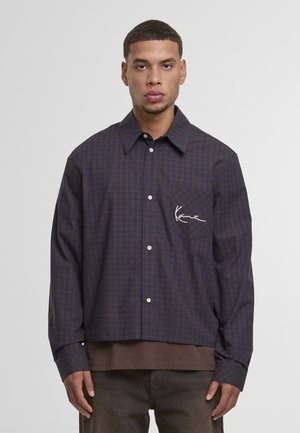 Muž nosiaci námornícky modrú a hnedú košeľu na zapínanie so štvorcovým vzorom a náprsným vreckom s bielym kurzívnym textom, pod ktorou má hnedé tričko.