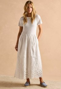 Weißes Baumwoll-Maxikleid mit kurzen Ärmeln und geraffter Taille, mit floralem Lochmuster entlang des Saums. Getragen mit blauen Espadrilles.