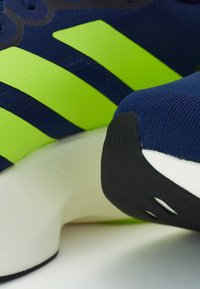 Pantofii sport cu partea superioară de culoare bleumarin, trei dungi îndrăznețe de un verde neon, material texturat și o talpă intermediară albă cu o talpă exterioară din cauciuc negru.