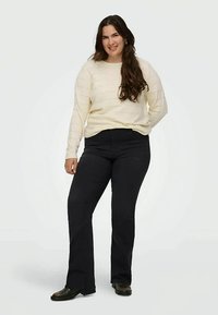 Jovem mulher a sorrir, de pé com uma mão atrás das costas, vestindo um suéter creme, calças pretas e sapatos pretos sobre fundo branco.