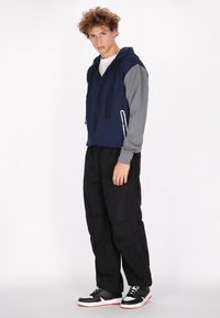 Giacca a cappuccio blu e grigia con tasca a zip, maniche grigie, abbinata a pantaloni neri e sneakers nere con accenti bianchi e rossi.