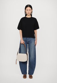 T-shirt noir oversize, jean baggy bleu taille haute, et sac bandoulière crème avec un motif géométrique et une sangle brune. Chaussures à plateforme marron.