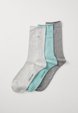 Tre sammenbrettede Calvin Klein sokker i lys grå, mintgrønn og mørk grå, ordnet overlappende på en hvit bakgrunn.