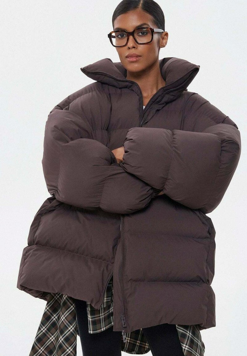 Smeđa pufasta jakna s oversized siluetom, visokim ovratnikom i prošivenom teksturom. Ima prednji patentni zatvarač i velike, mekane rukave.