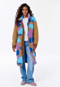 Kobieta z długimi kręconymi włosami, ubrana w długi beżowy płaszcz z niebieskimi i fioletowymi łatami, białą koszulę, niebieskie dżinsy i różowe trampki, stoi.