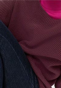 Suéter tejido burdeos con un patrón texturizado, superpuesto a una blusa rosa, combinado con denim oscuro. Se puede notar el detalle del suave tejido acanalado.