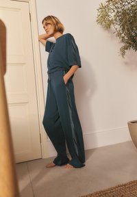 Tirkīza krāsa loungewear komplekts, kas sastāv no brīvā piegriezuma topa ar īsām piedurknēm un biksēm ar elastīgu jostu, kurām ir satīna sānu paneļi. Fons ar neitrālām krāsām.