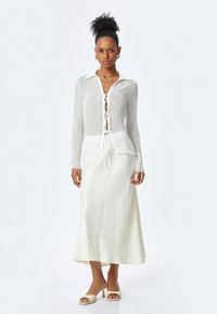 Chemise blanche légère à manches longues avec une ouverture nouée à l'avant, associée à une jupe en satin blanc fluide. Texture lisse, design superposé.