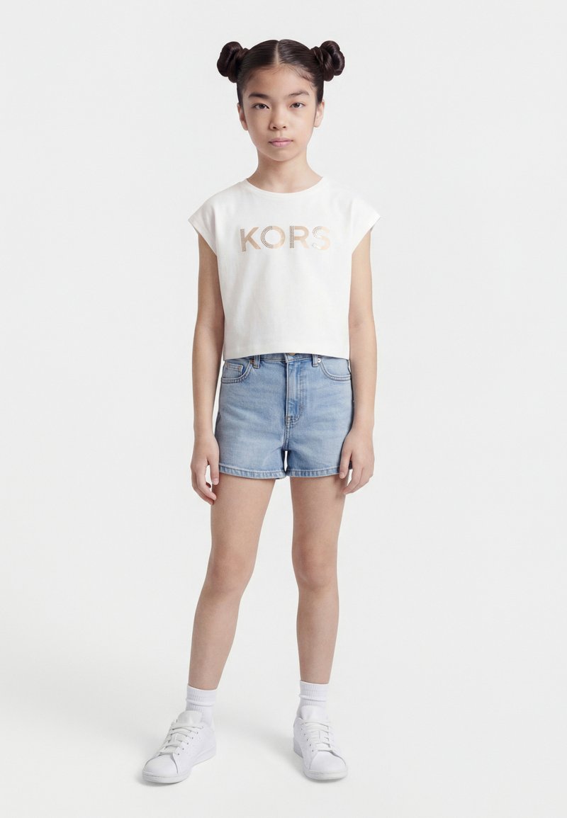 Κορίτσι όρθιο, φοράει λευκό κοντό αμάνικο τοπ με λογότυπο "KORS", ανοιχτό μπλε τζιν σορτσάκι, λευκές κάλτσες και λευκά αθλητικά παπούτσια.