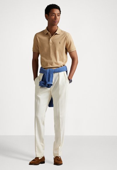 Homem vestindo uma camisa polo bege, calças brancas pregueadas, mocassins castanhos e um suéter azul amarrado à cintura, em pé com as mãos nos bolsos.