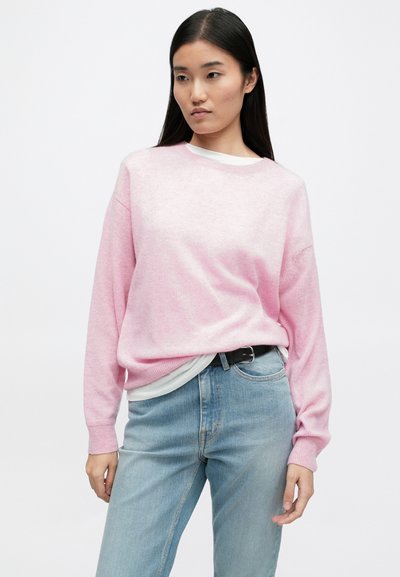 Sweater oversized rosa claro com mangas longas, punhos canelados, decote redondo, sobre uma camisa branca, combinado com jeans azul claro.