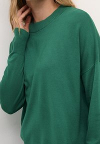 Zielony sweter z dzianiny z okrągłym dekoltem. Posiada długie, luźne rękawy i miękką teksturę. Prosty design, bez wzorów czy zdobień.