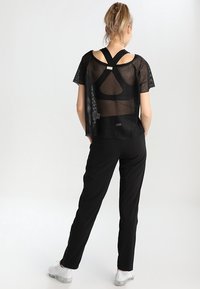 Top in rete nero con maniche corte, caratterizzato da un design incrociato sulla schiena, abbinato a pantaloni neri aderenti. L'outfit ha un aspetto sportivo.