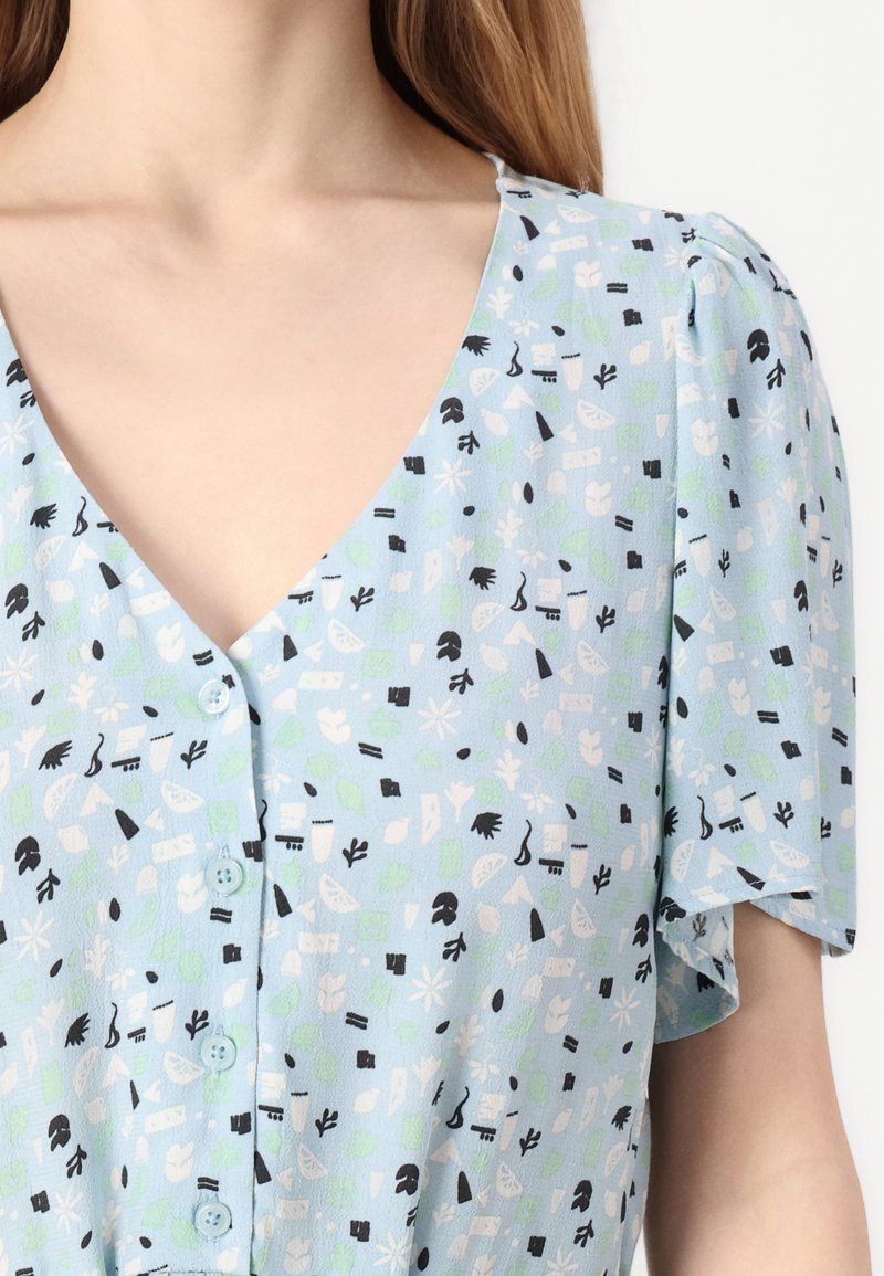 Jasnoniebieska bluzka z dekoltem w kształcie litery V, krótkimi rękawami i z przodu zapinaną na guziki. Posiada wzorzysty design z kształtami w kolorze zielonym, czarnym i białym.