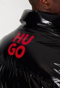 Czarna kurtka puchowa o błyszczącym wykończeniu, z wysokim kołnierzem i dużym czerwonym napisem "HUGO" na plecach. Widoczne fakturowa powierzchnia i detale szwów.
