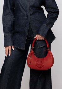 Red leather handbag with a braided handle, hardware accents, and an embossed logo. Worn by a person in dark denim clothing.