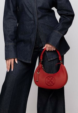 Red leather handbag with a braided handle, hardware accents, and an embossed logo. Worn by a person in dark denim clothing.