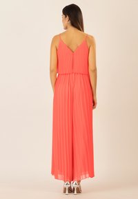 Mono de tirantes en coral con pliegues y espalda en V, que presenta un diseño fluido y zapatos de tacón alto en plateado.