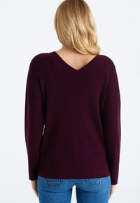 Nainen, jolla on vaaleat hiukset, pukeutuneena tumman violetin väriseen V-kaula-aukkosvettiin ja sinisiin farkkuihin, nähtynä takaa yksinkertaista valkoista taustaa vasten.