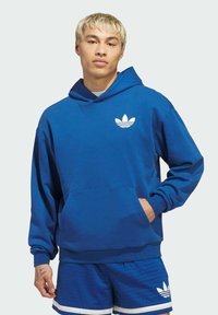 Niebieska bawełniana bluza z kapturem, z przednią kieszenią i białym akcentem logo. Noszona z niebieskimi szortami z podobnym logo i białym wykończeniem.