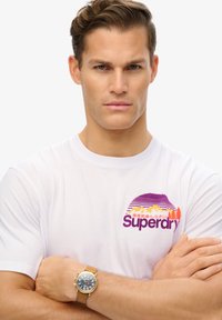 Bílý bavlněný tričko s fialovou a oranžovou grafikou hor a stromů, značky "Superdry." Hnědý kožený náramkový hodinky na zápěstí.