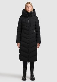 Kobieta ubrana w długą, czarną, pikowaną kurtkę puchową z dużym kapturem, czarne legginsy i czarne botki za kostkę, stojąca na jednolitym tle.