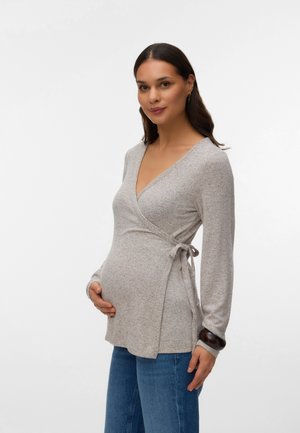 Schwangere Frau mit langen dunklen Haaren, die einen hellgrauen Wickelpullover und blaue Jeans trägt, hält sanft ihren Bauch und lächelt.