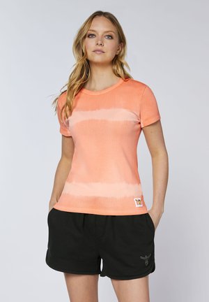 Young woman with long blonde hair wearing a short-sleeve orange tie-dye shirt and black shorts standing with hands in pockets.