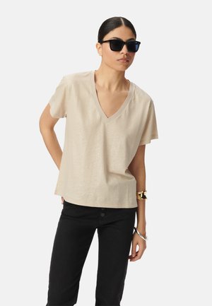 Beige V-Ausschnitt T-Shirt aus weichem Stoff, mit kurzen Ärmeln und lässiger Passform, kombiniert mit schwarzen Jeans und auffälligem Schmuck.