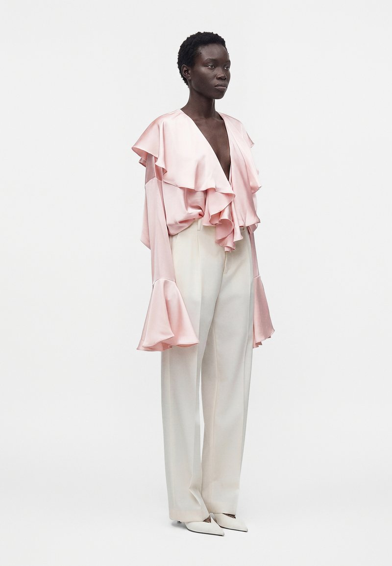 Mannequin portant un chemisier en satin rose pâle avec un décolleté en V profond et des manches à volants, associé à un pantalon blanc taille haute à jambes larges et à des chaussures blanches pointues.