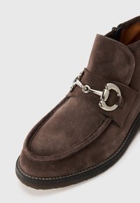 Zapato estilo slip-on de ante marrón con acentos de metal plateado, puntera redondeada y suela negra contrastante. Presenta detalles de costura y un acabado mate.