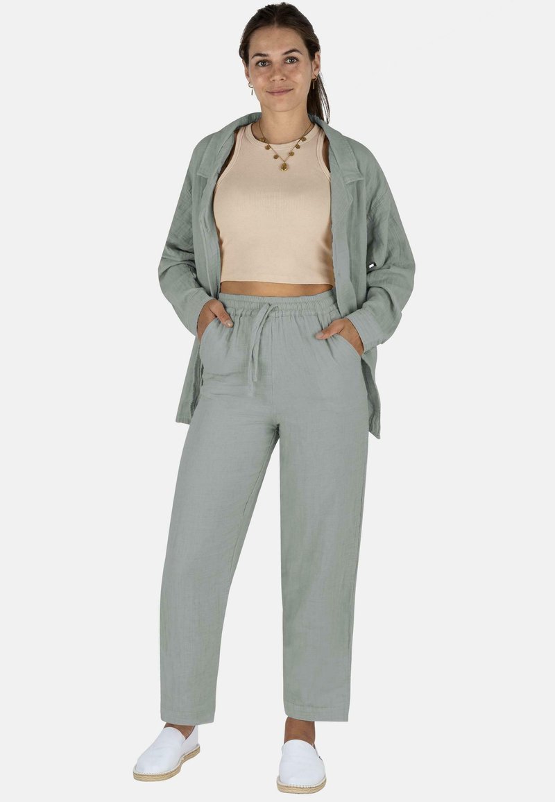 Leinenoutfit mit einem hellgrünen Langarmhemd, beigem Crop-Top und passenden Jogginghosen mit Kordelzug, ergänzt durch weiße Slippers.