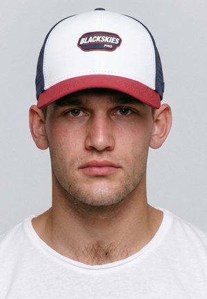 Junger Mann mit kurzen dunklen Haaren, der eine weiße und rote Blackskies Pro Cap und ein weißes Rundhalsausschnitt-Shirt trägt und geradeaus schaut.
