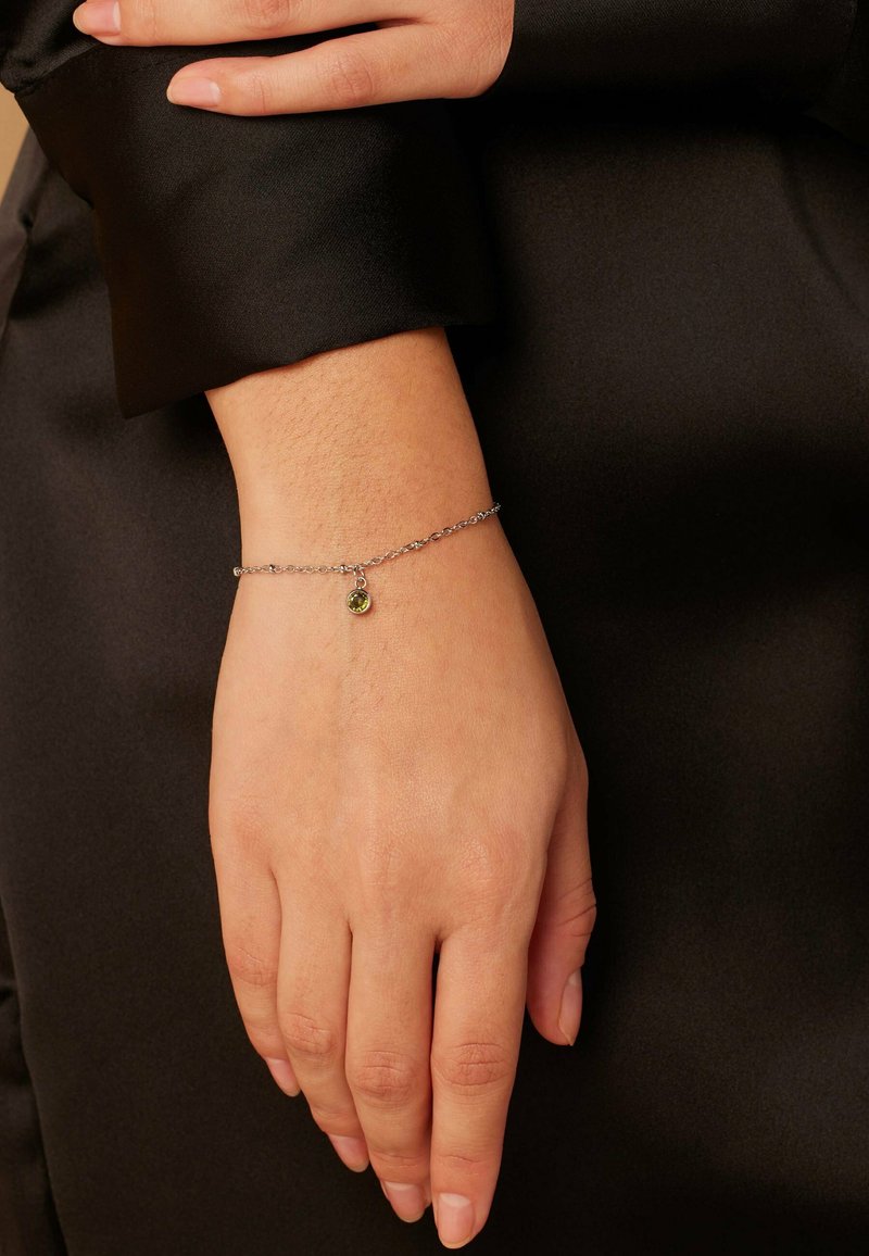 Zilveren kettingarmband met een kleine groene edelsteen hanger, gedragen op een hand met een natuurlijke nagellakafwerking tegen een zwarte stoffen achtergrond.