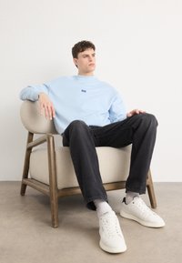 Felpa azzurra con un piccolo logo, jeans neri e sneakers bianche. Seduto su una sedia beige con braccioli curvi e gambe in legno.