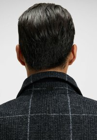 Cappotto a quadri scuri con un misto di lana textured, caratterizzato da un colletto a punta e un piccolo etichetta nera sulla parte posteriore.