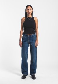 Black sleeveless top, blue high-waisted jeans with a straight leg, and black slip-on shoes. Three layered necklaces and visible tattoos.