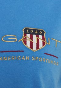 Modré bavlněné tričko s vyšitou šerpou se symbolem a červenými pruhy a zlatým textem, který zní "GANT AMERICAN SPORTSWEAR" a "1949."