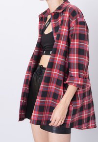 Rød og sort ternet skjorte med krave, der har opsmøgede ærmer og knaplukninger. Båret over en sort crop top og sorte shorts.