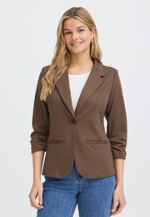 Kvinde med langt blond hår iført en brun blazer over en hvid top og blå jeans, smilende mod en ensartet lys baggrund.