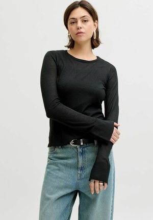 Jeune femme aux cheveux courts et bruns portant de grandes boucles d'oreilles en anneau, un haut noir à manches longues et un jean bleu clair, sur un fond uni.