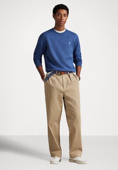 Zils kokvilnas džemperis ar apaļu kakla izgriezumu, kombinēts ar gaisīgi bēšām biksēm ar krokām un svītrainu jostu. Izskatu papildina balti sporta apavi.