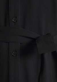 Chemise en tissu noir avec boutonnière et ceinture à nouer. La texture est lisse, avec un col classique et une coupe ajustée.